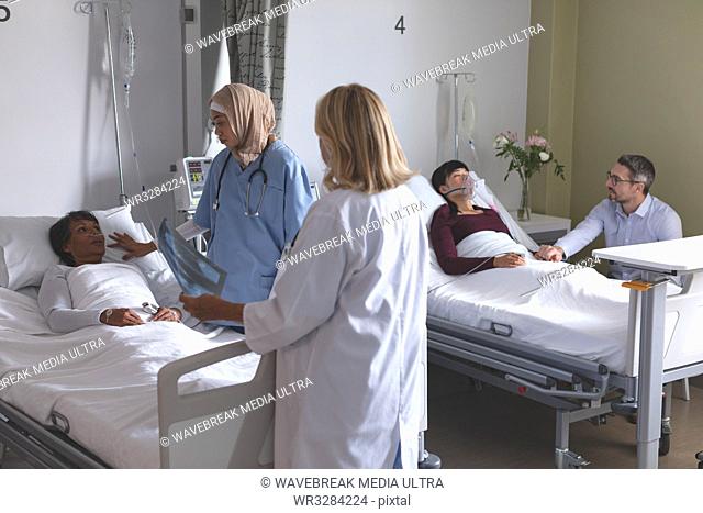 Side view of diverse female doctors interacting with female patient in the ward at hospital. In the background Caucasian man is holding the hand of Asian woman...