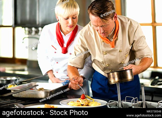 Two chefs in teamwork - man and woman - in a restaurant or hotel kitchen cooking delicious food, he is decorating the dishes, she is watching