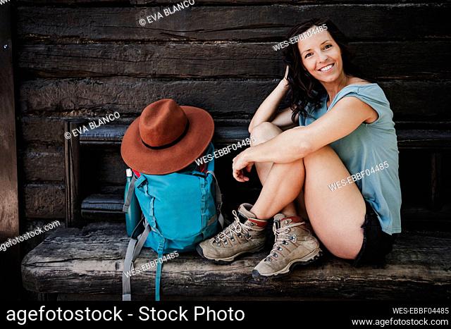 Happy woman with hand in hair sitting by backpack on bench outside cottage