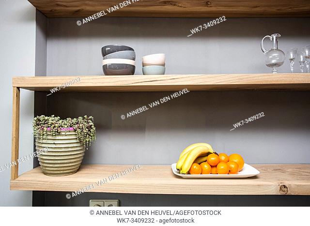 Shelf with various things and fruits on the table, fresh bananas and orange healthy snack. green plant and glasses grey wall