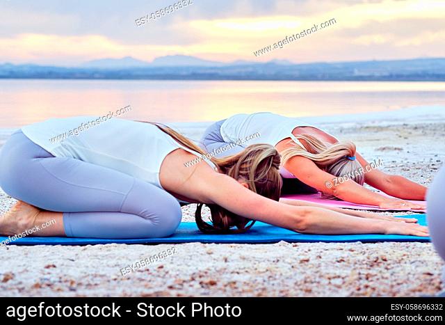 Women wearing sportswear practising yoga in nature during sunset sunrise on lake sea coast, yogi do Child pose Balasana exercise, working out outdoors