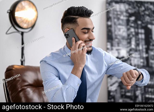 Punctuality. Young bearded business man smiling talking on smartphone looking at wrist watch in bright room