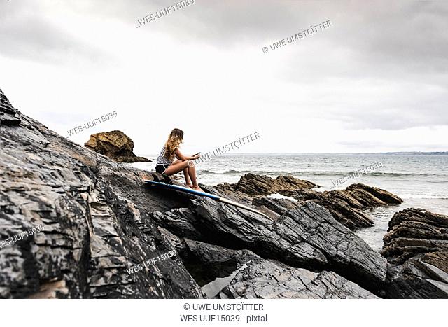 Young woman with surfboard sitting on the beach, using smartphone