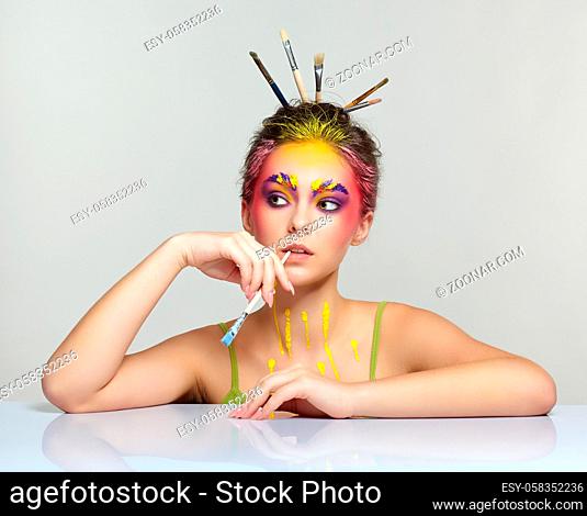 Portrait of young woman posing at the table with brushes in hair and in her teeth. Unusual female art make-up with paint on brows, hair and around eyes