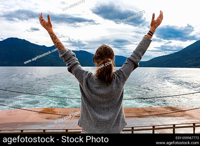 Close up from behind of a young lady on the deck of a passenger ferryboat. Arms raised in enthusiastic appreciation of the landscape. Freedom concept