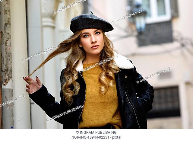 Blonde russian woman in urban background. Beautiful young girl wearing beret, black leather jacket and mini skirt standing in the street