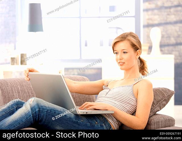 Young woman lying on sofa at home, browsing internet on laptop. Looking at screen, smiling
