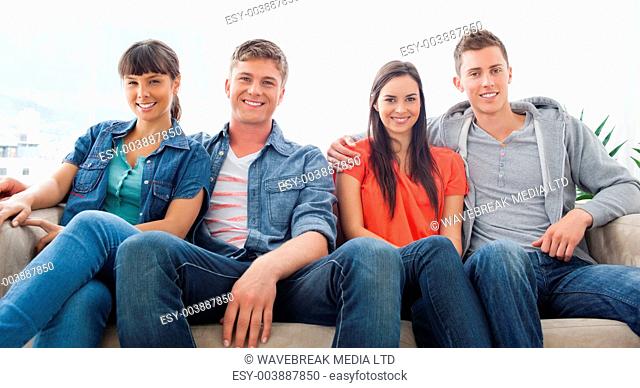 Two smiling couples sit on the couch beside one another as they look forward into the camera