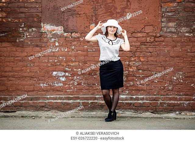 Photo of a woman in a white hat, blouse and black skirt, standing against the backdrop of an old vintage brown brick wall