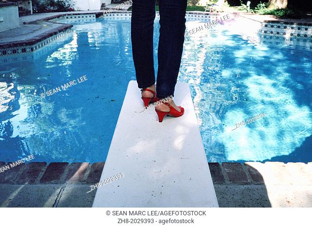 A woman wearing bright red high heel shoes on a diving board in front of a swimming pool