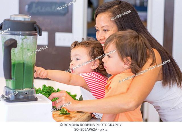 Mid adult woman making green smoothie for two toddlers in kitchen