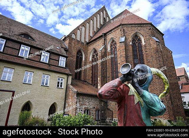 PRODUCTION - 20 July 2023, Brandenburg, Prenzlau: A harlequin figure sits making music in front of the former Dominican monastery in Prenzlau