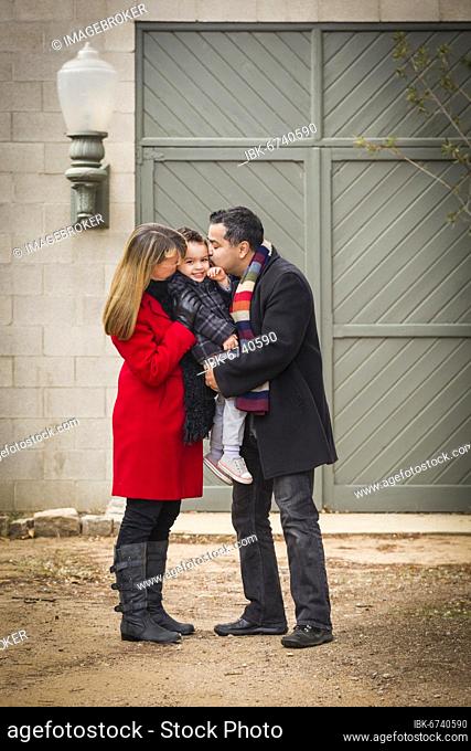 Young mixed-race couple in winter clothing hugging and kissing son in front of rustic building together