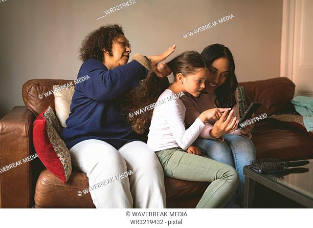Grandmother is making hairstyle of granddaughter while using mobile with her mother