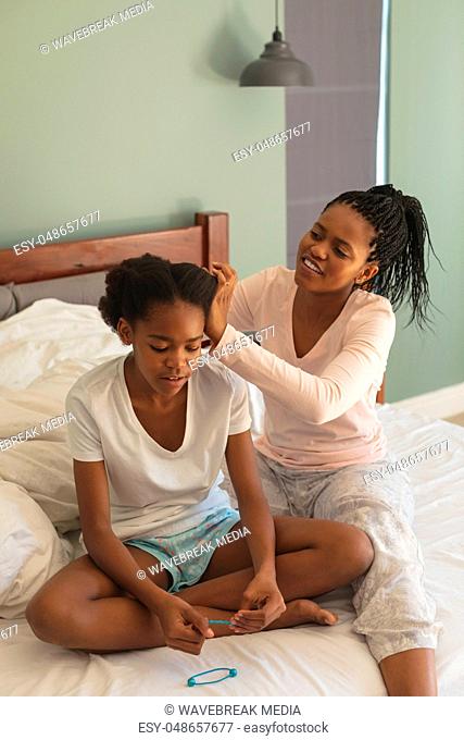 Happy African American mother hairdressing daughter on bed in bedroom