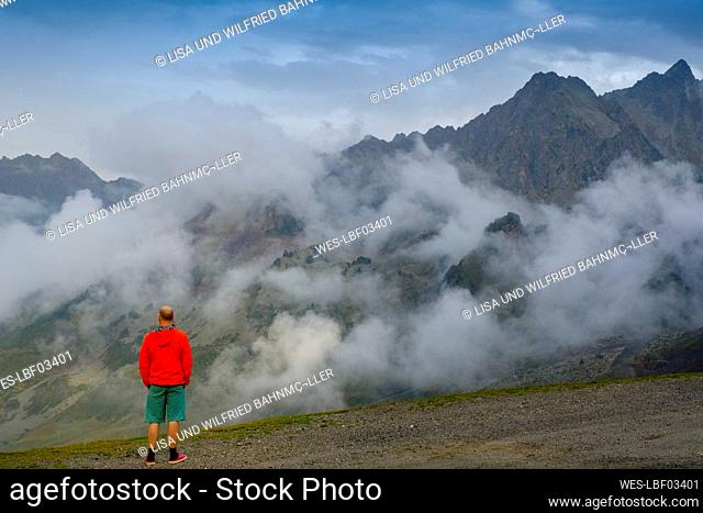 Man in vibrant red jacket looking at fog floating over Col du Tourmalet pass, France