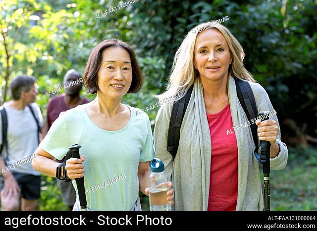 Two middle-aged women with hiking poles walking in the woods with their partners behind them