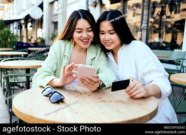Smiling women shopping online through smart phone at sidewalk cafe