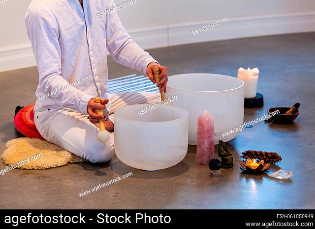 Man dressed in white, sitting on a sheep skin with a close up on his hands playing crystal bowls with sacred objects displayed like stones, candles and more