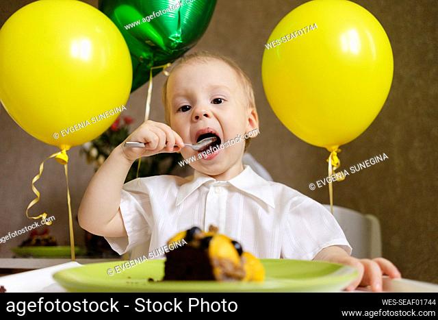 Boy eating cake with balloons behind at home