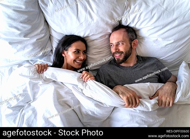 Couple looking at each other while lying on bed with blanket at home