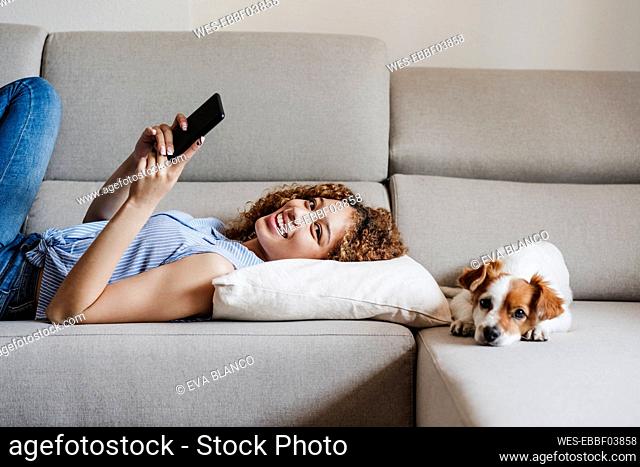 Smiling woman with mobile phone lying by Jack Russell Terrier on sofa at home