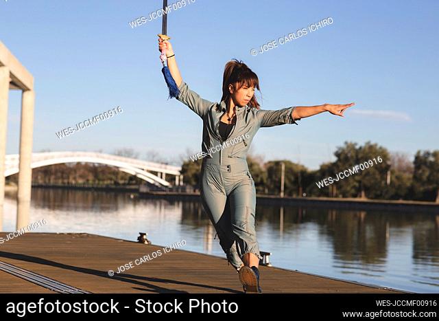 Young female athlete practicing martial arts with sword against lake in park on sunny day