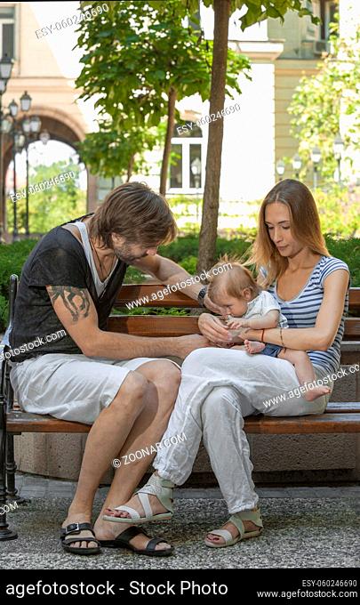 Happy young beautiful family of three is sitting on a bench outdoors in a european city