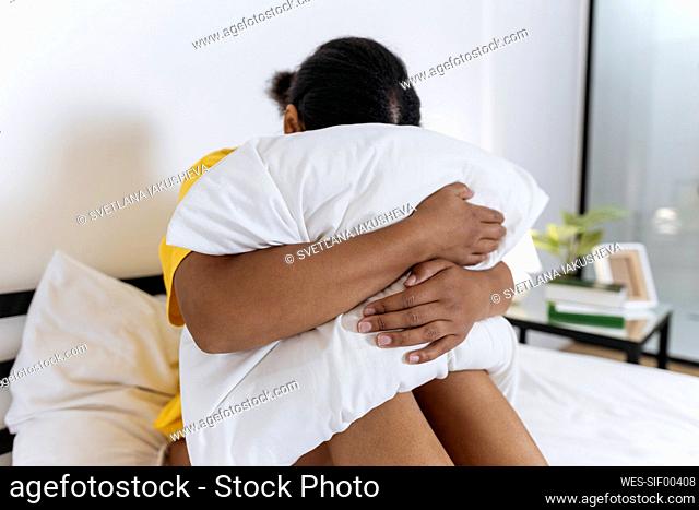 Upset woman hiding face in pillow while sitting on bed