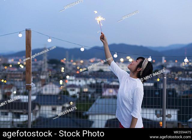 Smiling young Japanese woman holding sparkler on a rooftop in an urban setting