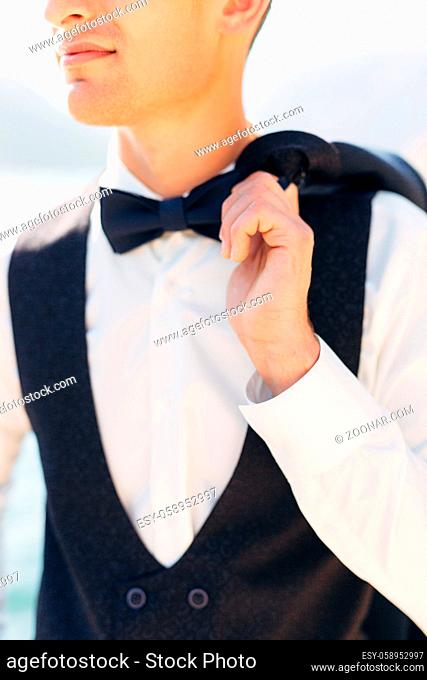 A man in a white shirt, vest and with a bow tie holds a jacket thrown over his shoulder . High quality photo