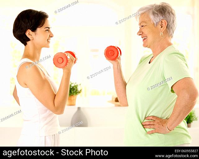 Healthy elderly woman doing dumbbell exercise with personal trainer at home, smiling