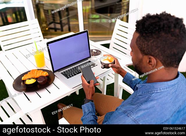 African american man sitting in cafe using laptop and smartphone drinking coffee eating breakfast
