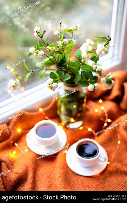 A white snowberry in a bouquet stands with two cups of coffee on the windowsill near the window, beautiful lights, bokeh in the window