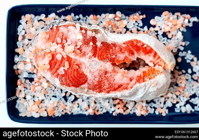 Fresh raw red salmon steak with large coarse pink salt prepared for baking on the grill lies on a blue plate. Healthy seafood food