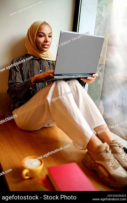 Arab girl in hijab sitting on windowsill and using laptop, university cafe interior on background. Muslim woman with books sitting in library