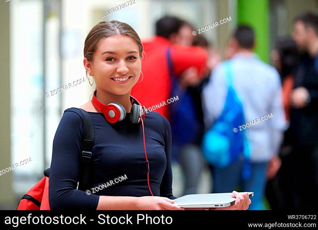 portrait of a happy young student getting ready for class while waiting in the college hallway with headphones and laptop