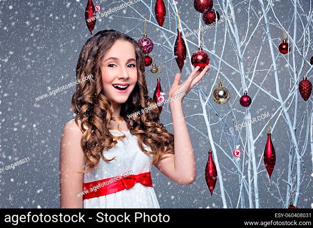 Beautiful teen girl with long curly hair in white dress with red ribbon belt standing near white tree branches wiht christmas decorations hanging on it