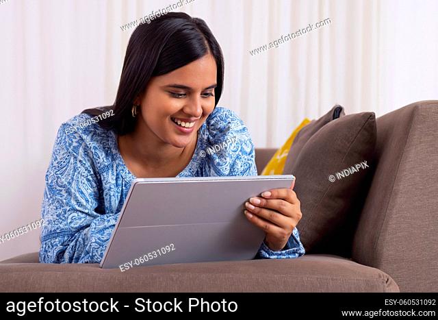 Beautiful Indian girl watching tablet while lying on sofa