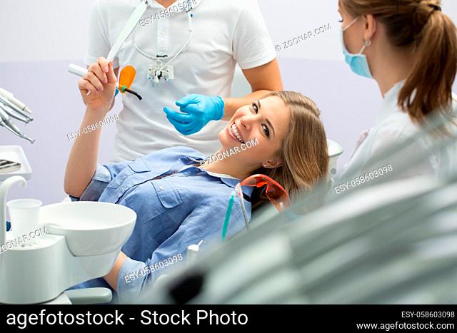 Young smiling girl in blue shirt with a mirror in the right hand on the patient chair in the dental cabinet. Next to her there is a dentist and a female...