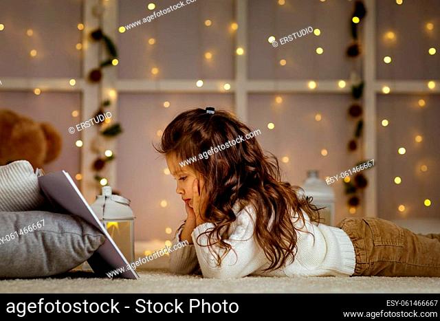 Happy curly little child girl is reading fairy tale book on the background with lights. Merry Christmas