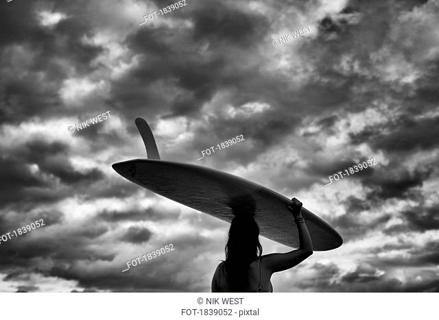Female surfer carrying long board overhead under cloudy sky