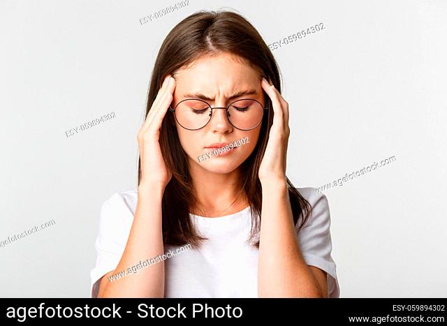 Close-up of young woman in glasses touching head and grimacing from pain, having headache or migraine, feeling sick