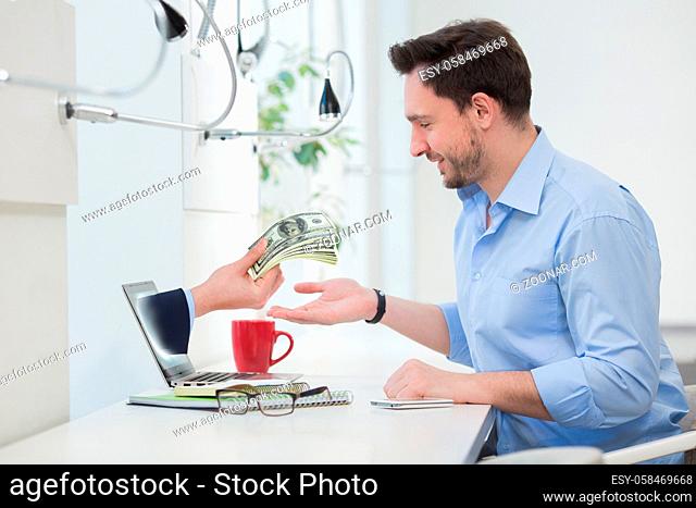 Happy businessman sitting in front of laptop computer while one hand holding hundreds of dollar bills through laptop screen concept for internet e-commerce
