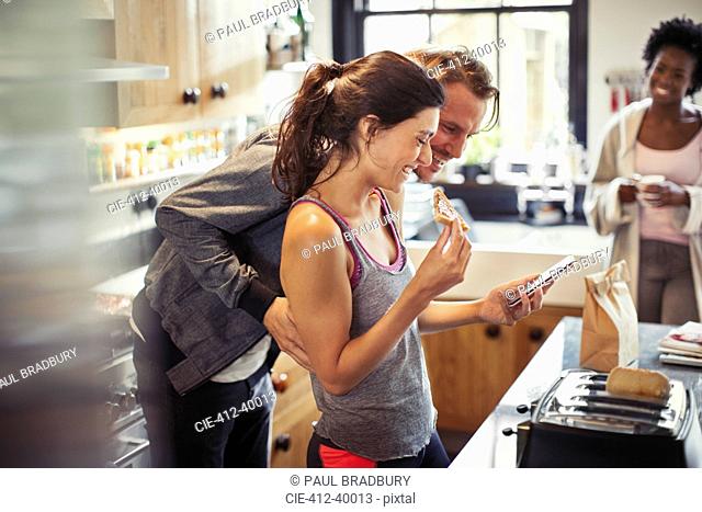 Smiling couple texting with smart phone, eating toast in kitchen