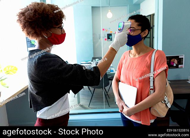 Mixed race woman wearing face mask checking temperature of female colleague