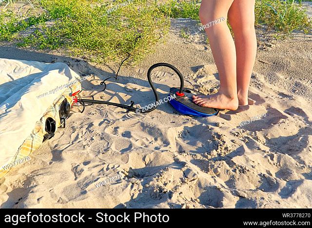 A woman with air foot pump pumps an inflatable mattress or air bed at sandy beach. Foot inflates air mattress with foot pump on sand