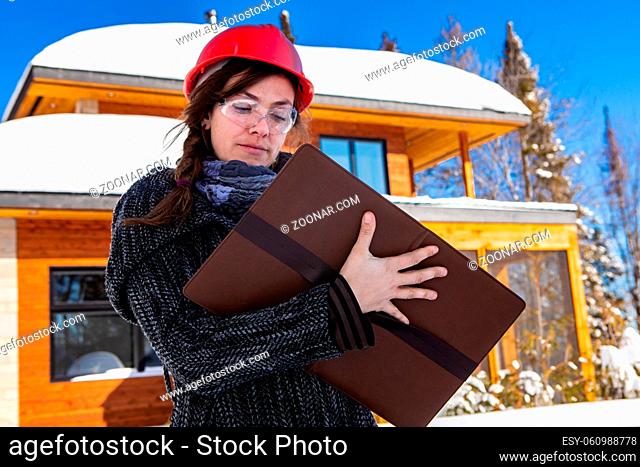 low angle view of a female civil engineer standing outdoor holding a clipboard and taking notes, the snow on wooden modern building in the background