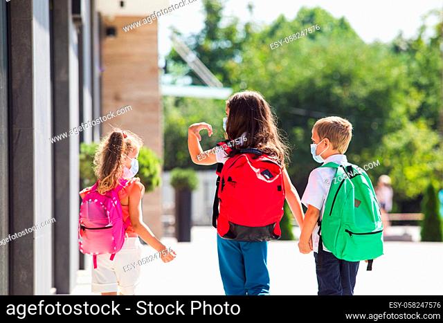 Back view of three schoolchildren who going back to school after pandemic over. Kids with protective masks and backpacks for safety