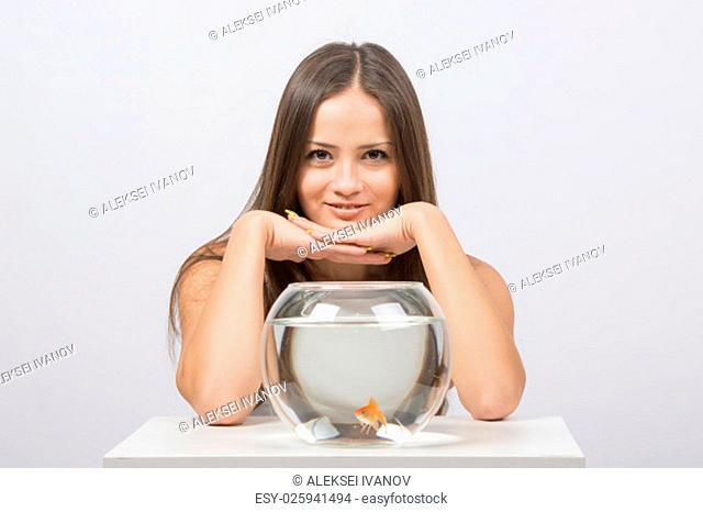 A young girl sits next to a round aquarium in which swimming goldfish
